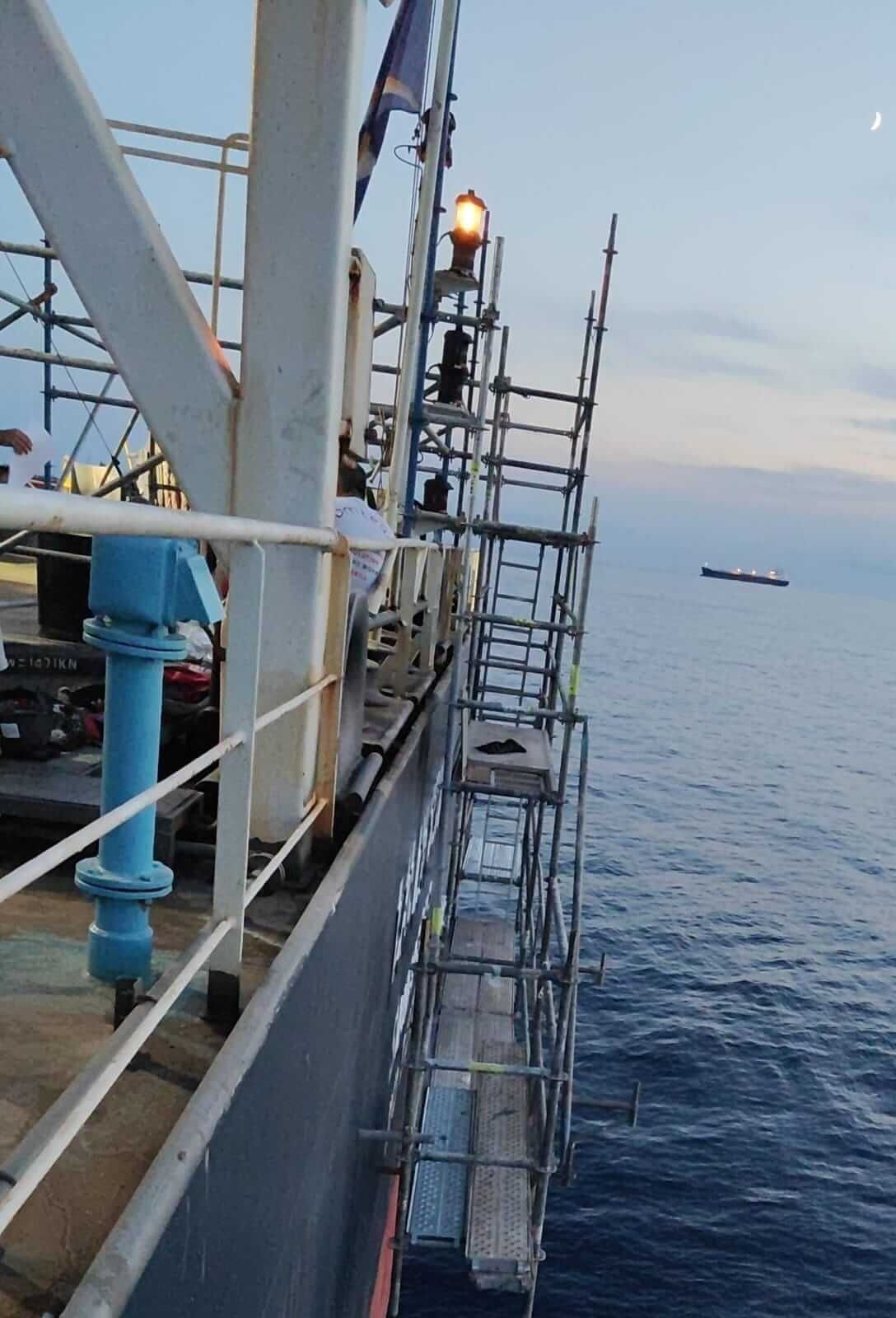 Andamio fijado al costado de un barco sobre el océano, con un barco a lo lejos y la luna visibles.