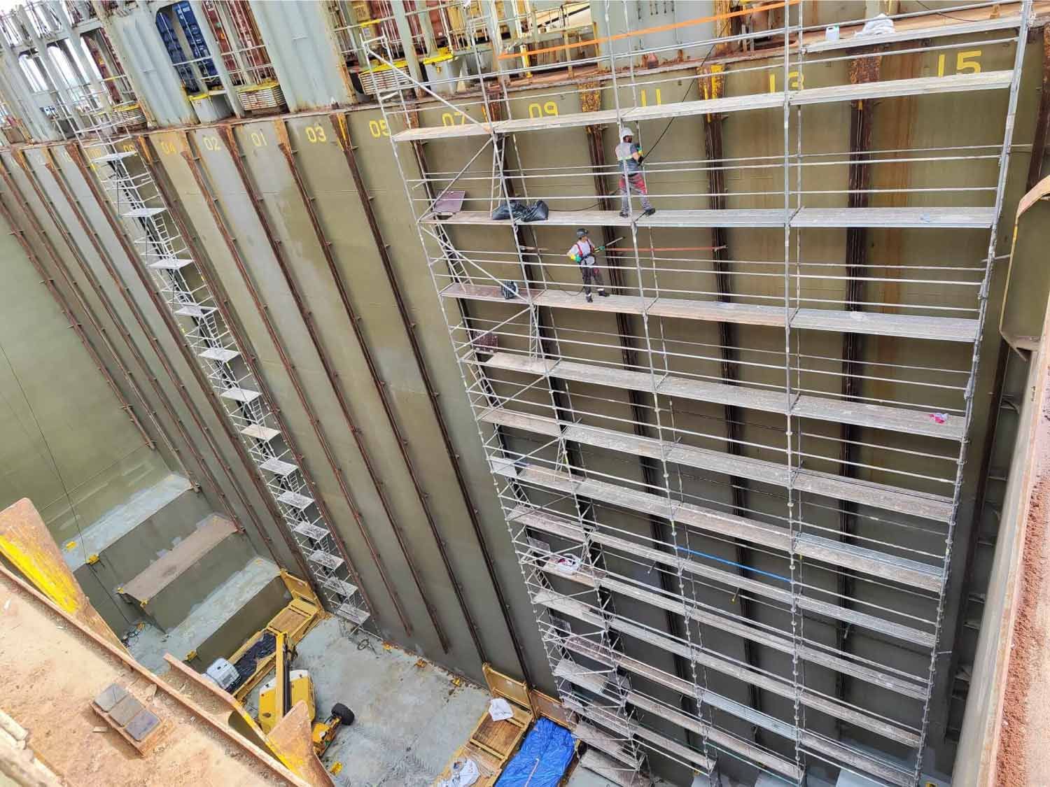 Trabajadores en andamios dentro de una gran estructura industrial. Paredes metálicas con paneles numerados.