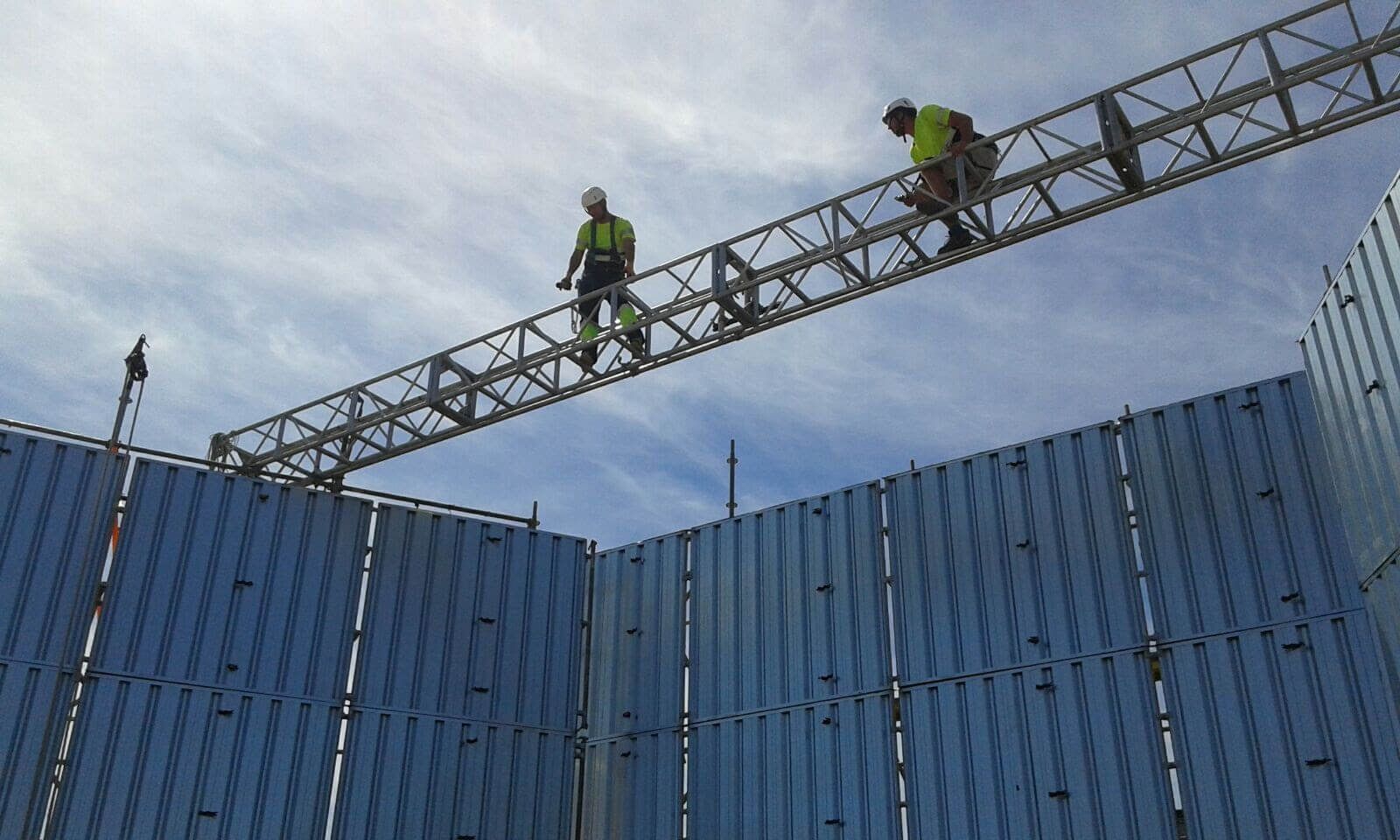 Dos trabajadores de la construcción en un andamio de metal sobre una pared corrugada azul bajo un cielo nublado.
