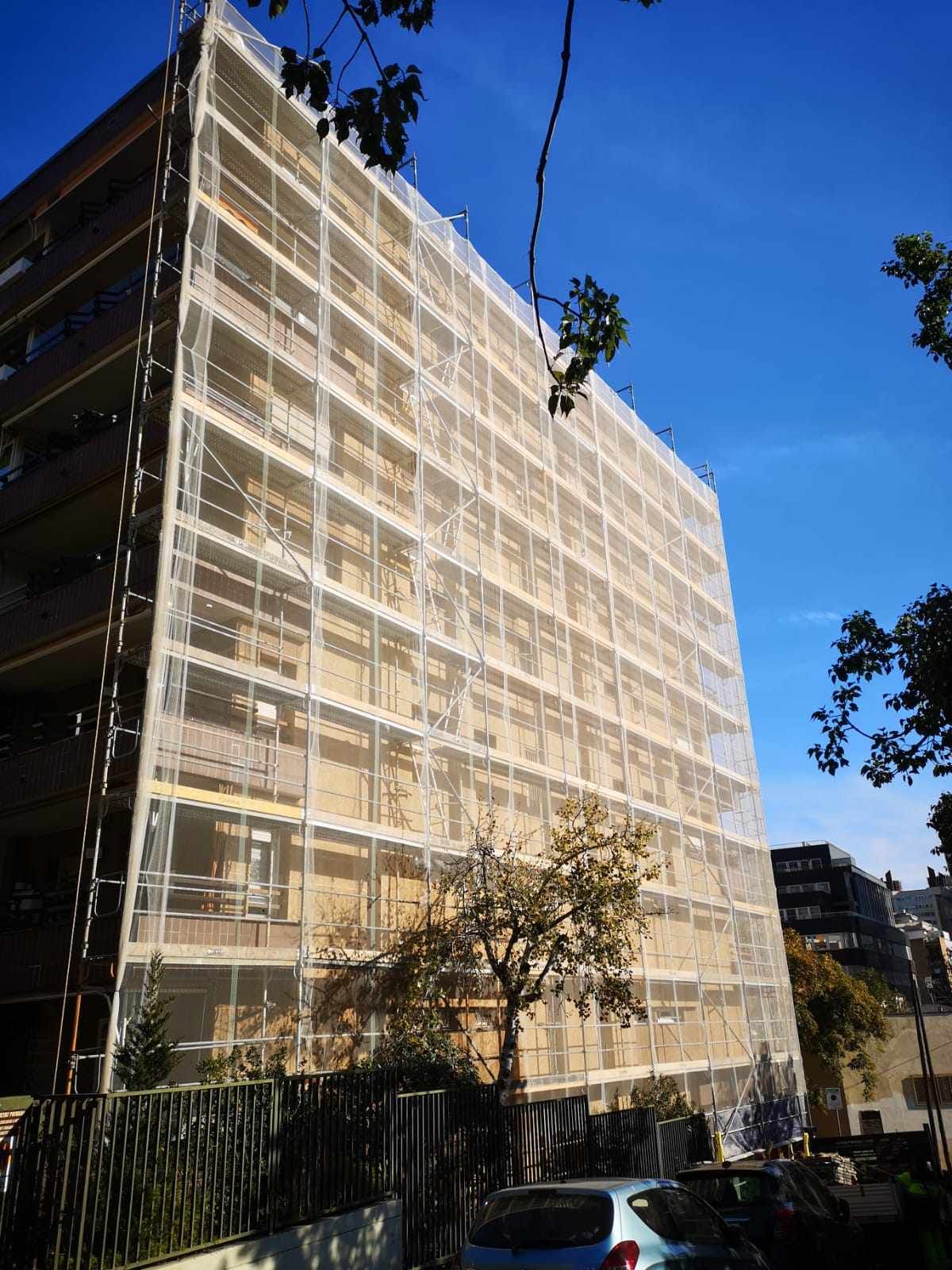 Edificio en construcción, cubierto con una red de andamios blanca contra un cielo azul, árboles en primer plano.