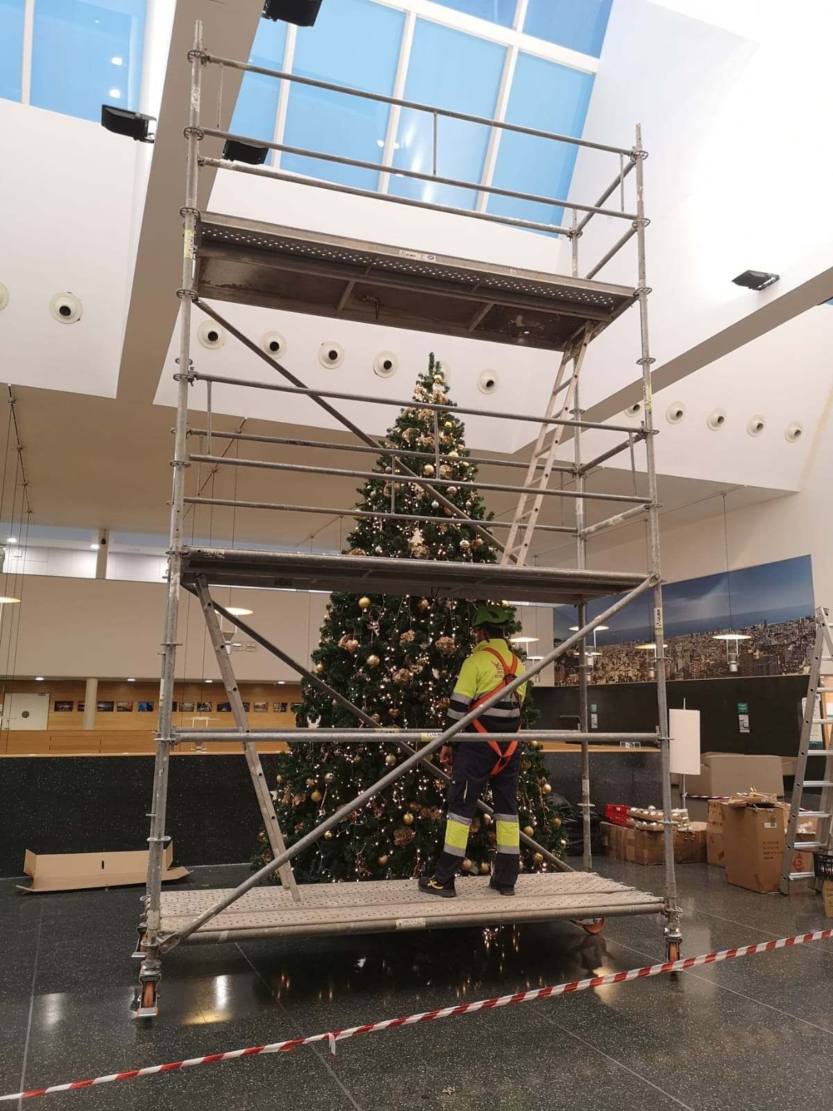 Andamio junto a un árbol de Navidad decorado dentro de un edificio. Una persona con ropa de trabajo se encuentra en la plataforma inferior.