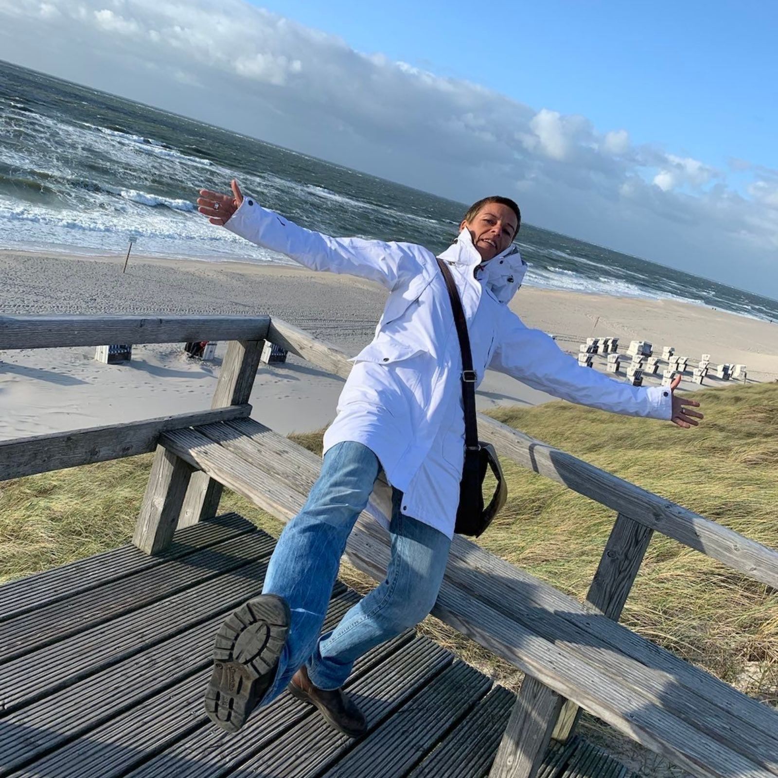 Eine Person in einem weißen Kittel und Jeans steht mit ausgestreckten Armen auf einem Holzdeck mit Blick auf den Ozean.