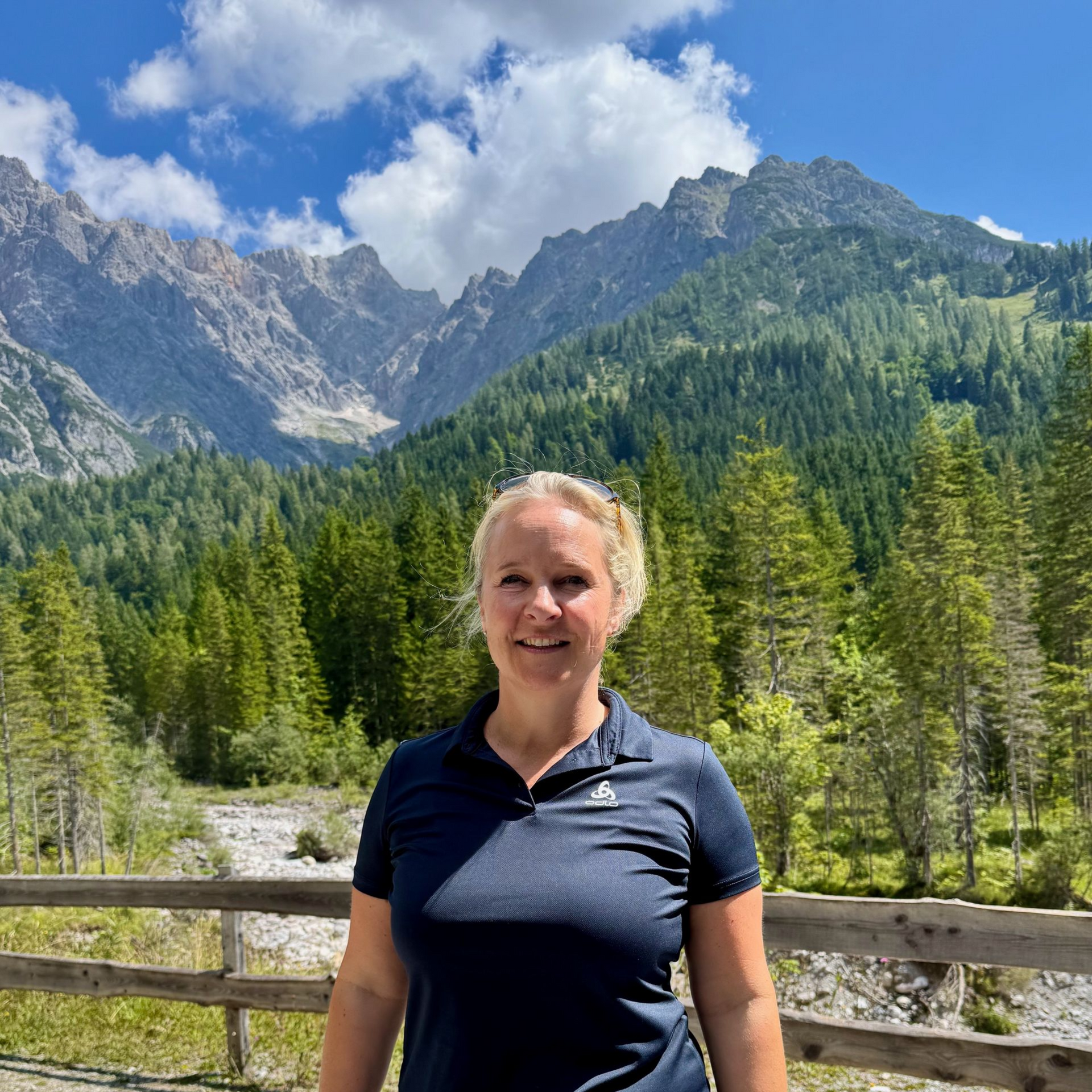 Frau vor Bergen und Bäumen, trägt ein blaues Hemd und lächelt.