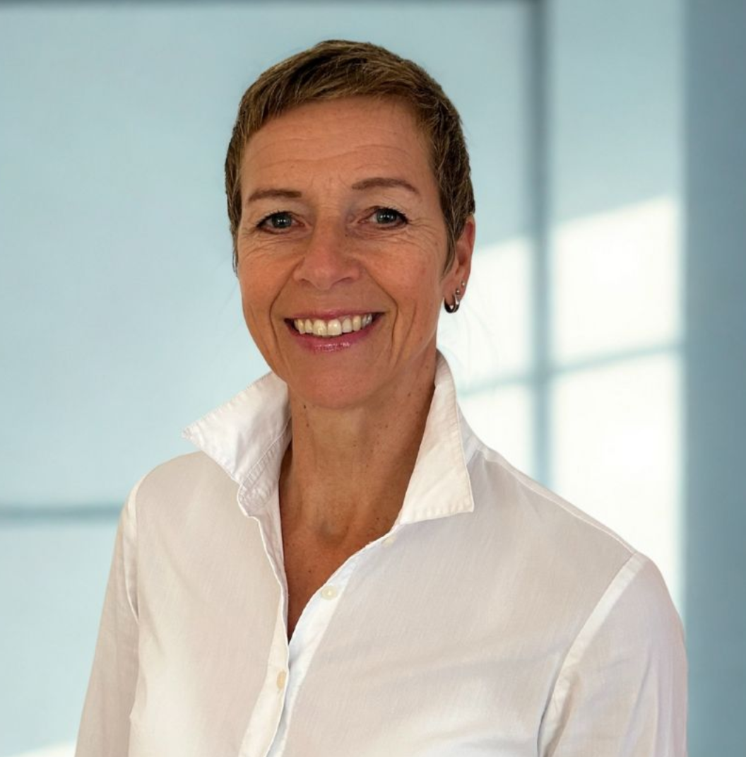 Kathrin Bertram Lächelnde Frau mit kurzen Haaren, trägt ein weißes Hemd mit Kragen und steht vor einem unscharfen Hintergrund.