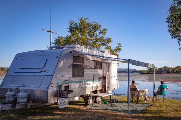 Caravane au bord d'un lac, deux personnes assises sous un auvent par une journée ensoleillée.