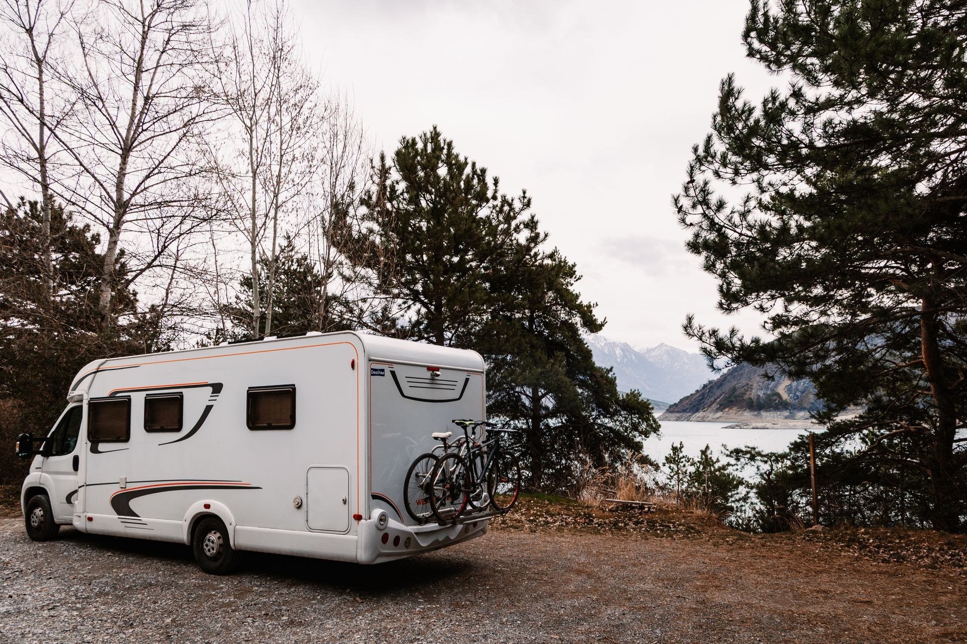 Un camping-car blanc garé au bord d'un lac boisé, avec des montagnes à l'horizon.