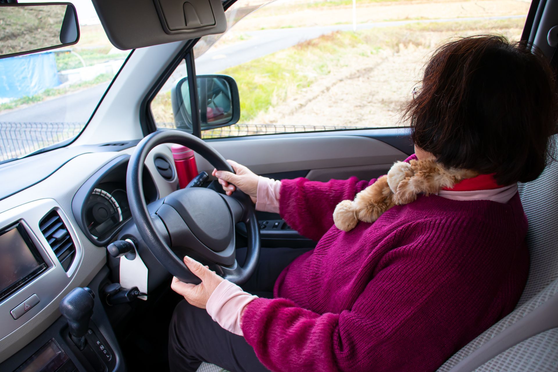 Un conducteur, tenant le volant de sa voiture, porte un pull et une écharpe magenta, sur une route de campagne.