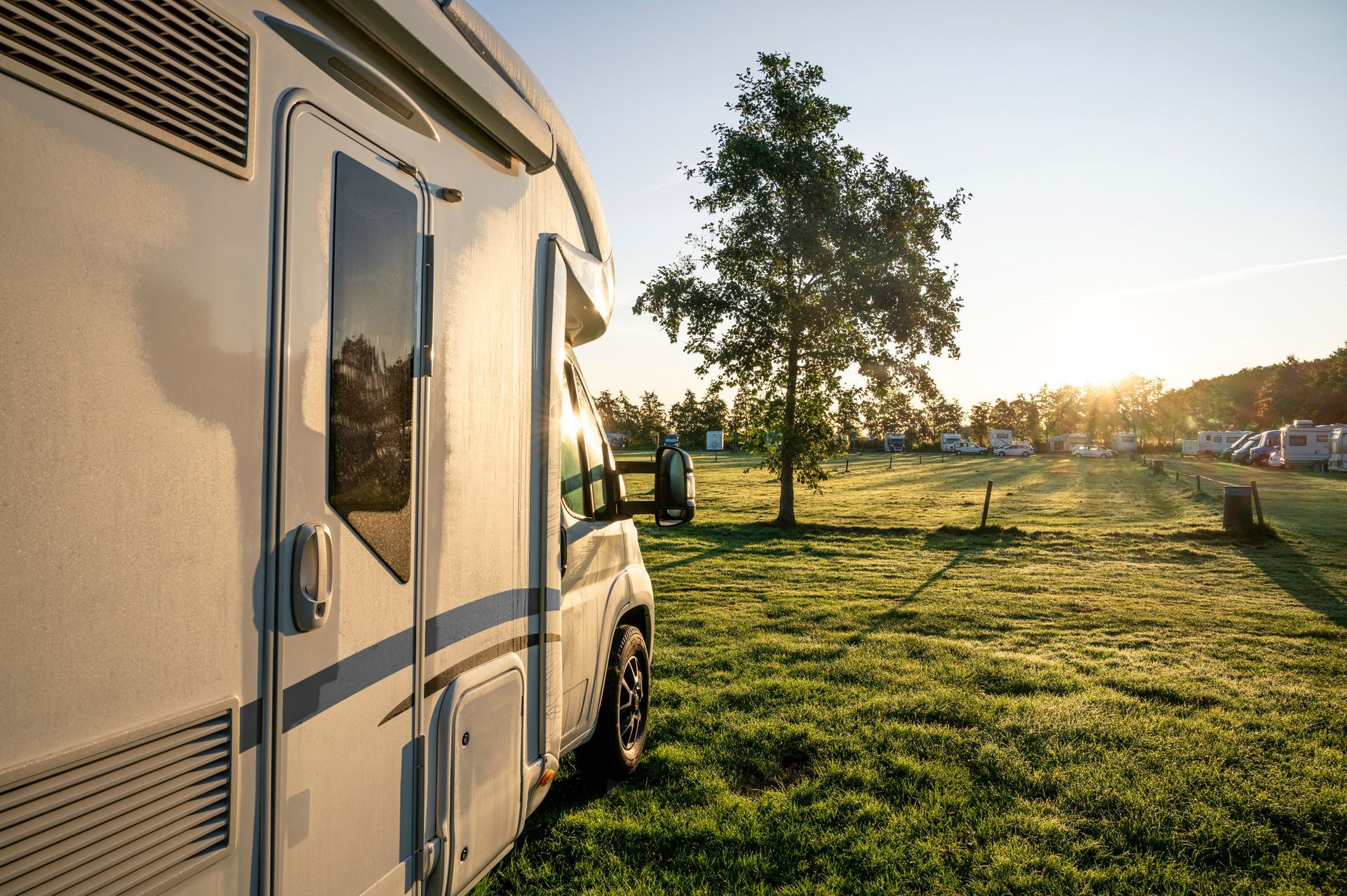 Un camping-car blanc est garé sur l'herbe au coucher du soleil, près d'un arbre et d'un champ.