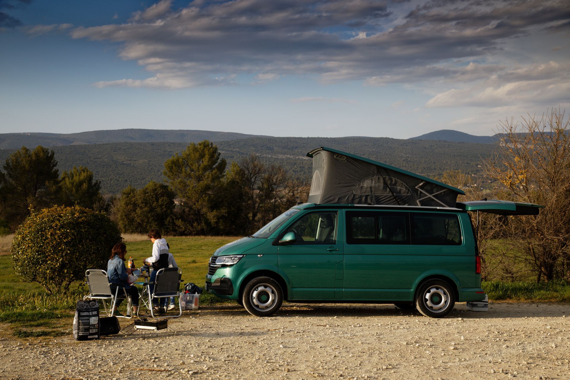 Un camping-car vert avec une tente de toit, à côté de campeurs profitant d'un point de vue panoramique sur les montagnes.