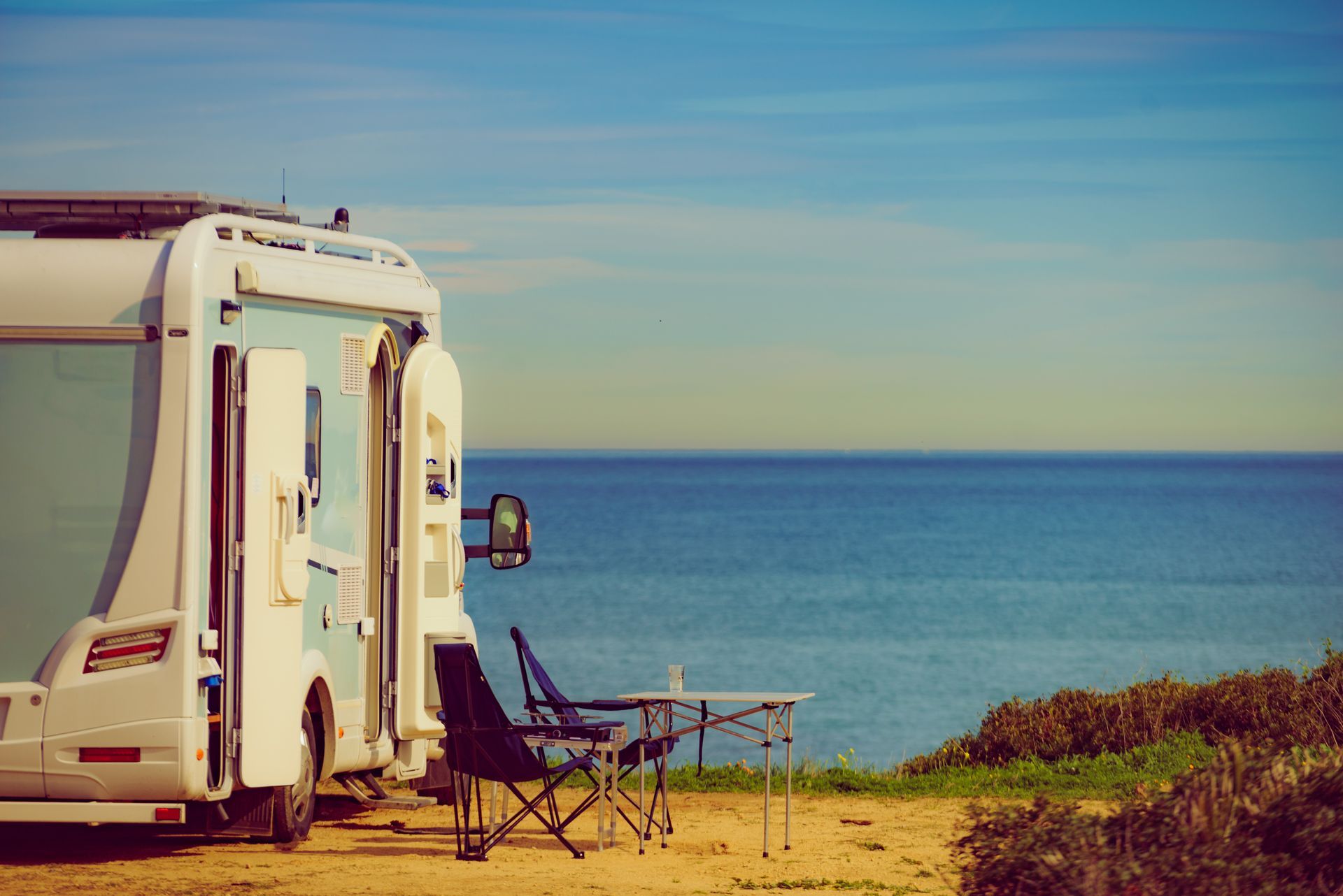 Un camping-car à côté d'une chaise sur une plage de sable, avec une mer bleue calme et un ciel dégagé en arrière-plan.