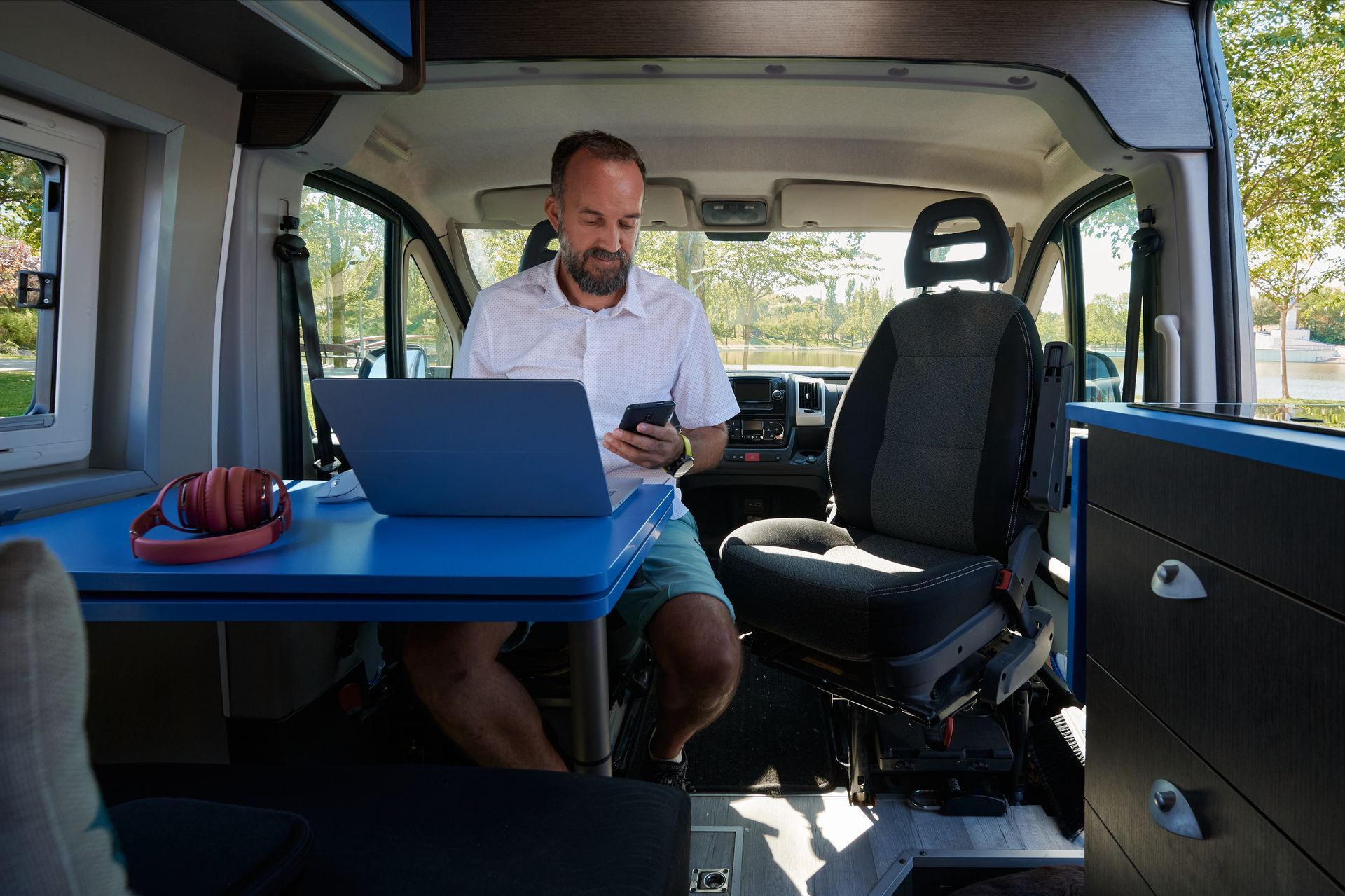Un homme utilise un ordinateur portable et un téléphone à l'intérieur d'un camping-car avec une table bleue et des sièges noirs.