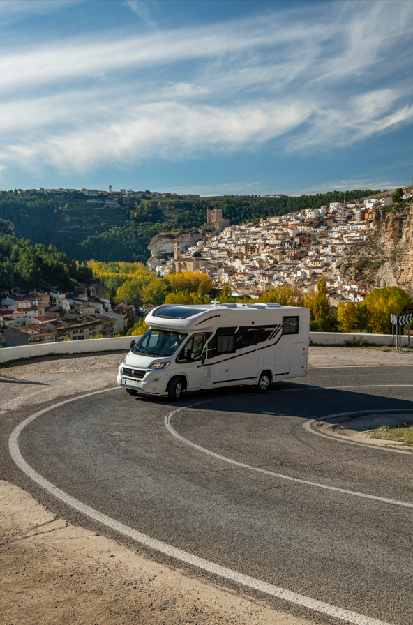 Un camping-car blanc sur une route de montagne sinueuse surplombant un village à flanc de colline sous un ciel bleu.