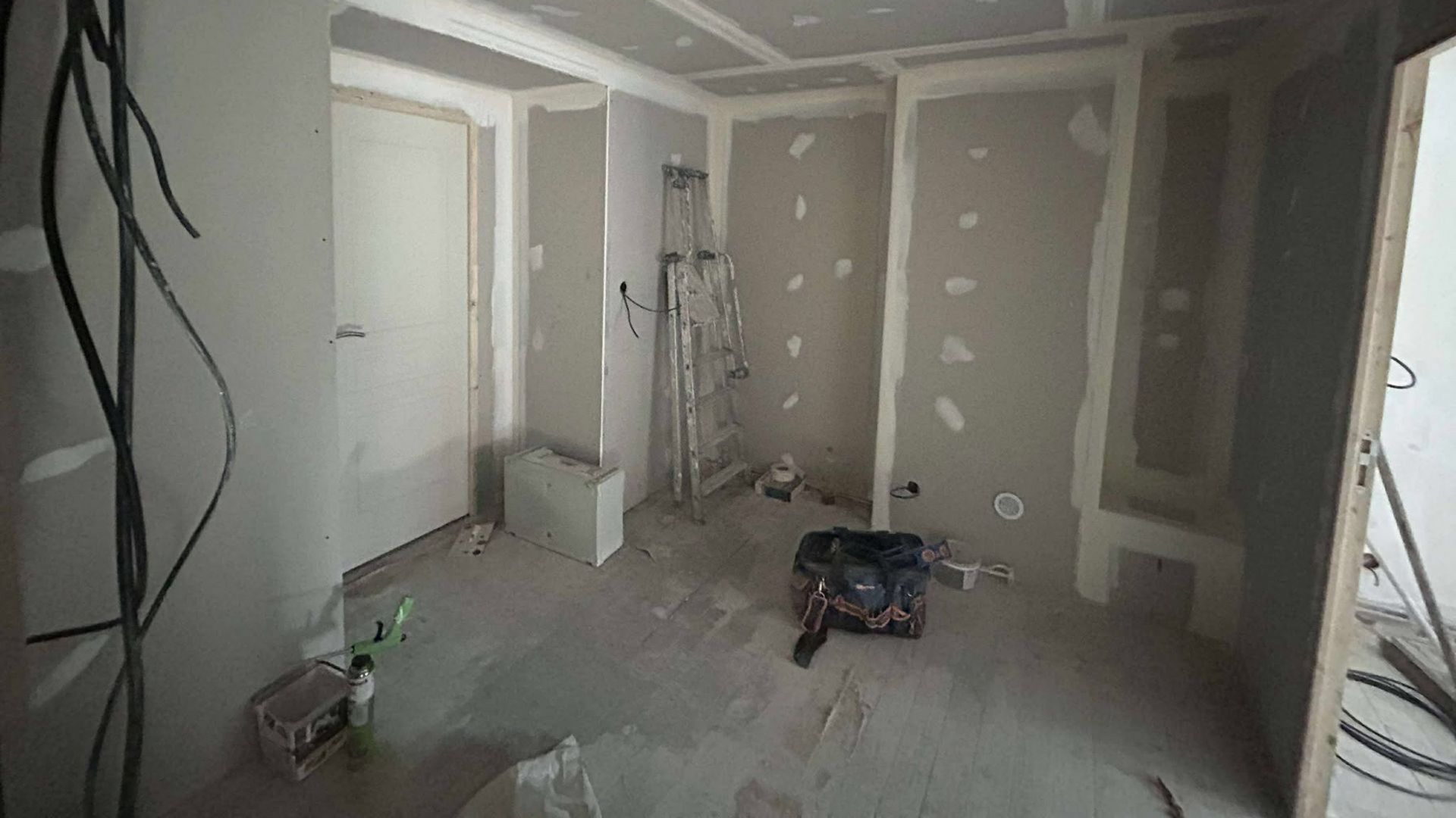 Pièce en travaux, murs non finis avec des plaques de plâtre, câbles et outils au sol.