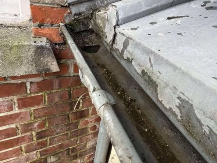 Gouttière métallique sale et obstruée, fixée à un bâtiment en briques.