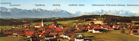 Panoramablick auf ein Dorf mit rotgedeckten Häusern, Kirchturm und grünen Hügeln unter klarem Himmel