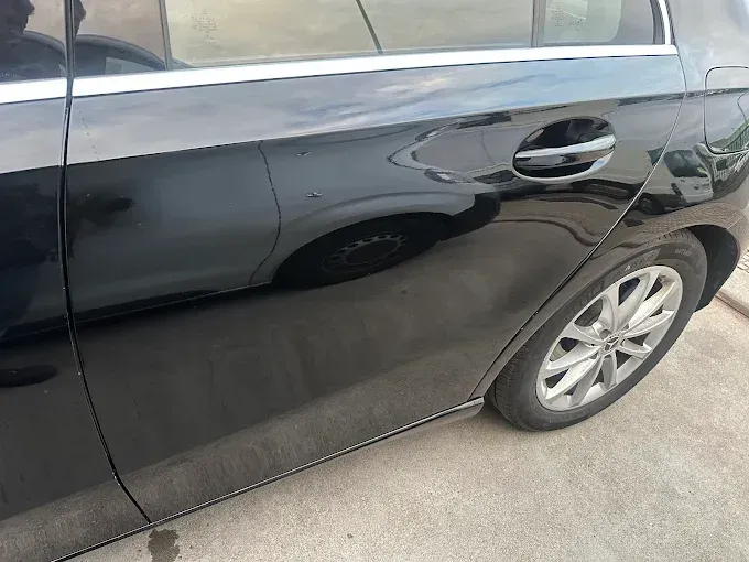 Lateral de un coche negro, manija de la puerta visible. Superficie reflectante que muestra el entorno. 