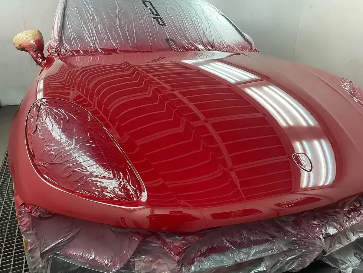 Capó de coche rojo recién pintado en taller, reflejos de luces sobre la superficie brillante.