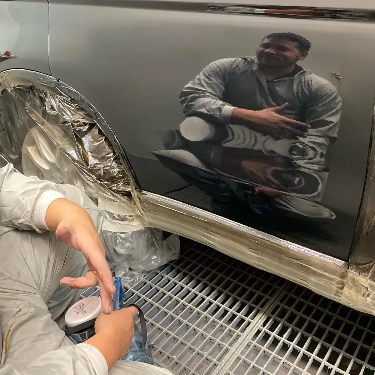 Dos personas trabajan en un coche negro; una se refleja. Llevan trajes claros y trabajan en un taller bien