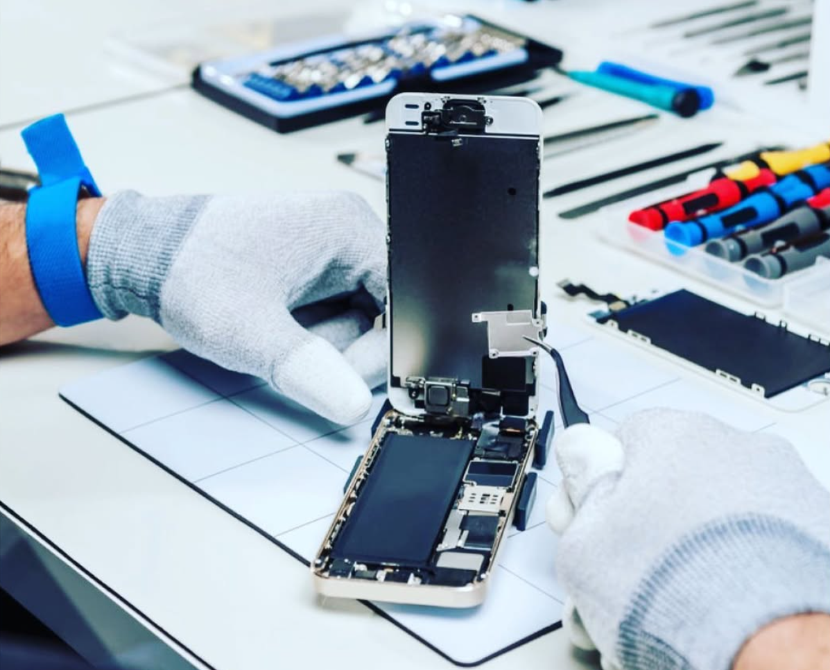 Eine Person zerlegt ein Smartphone mit einem Schraubenzieher auf einem Holztisch; Bauteile und Einzelteile liegen verstreut.