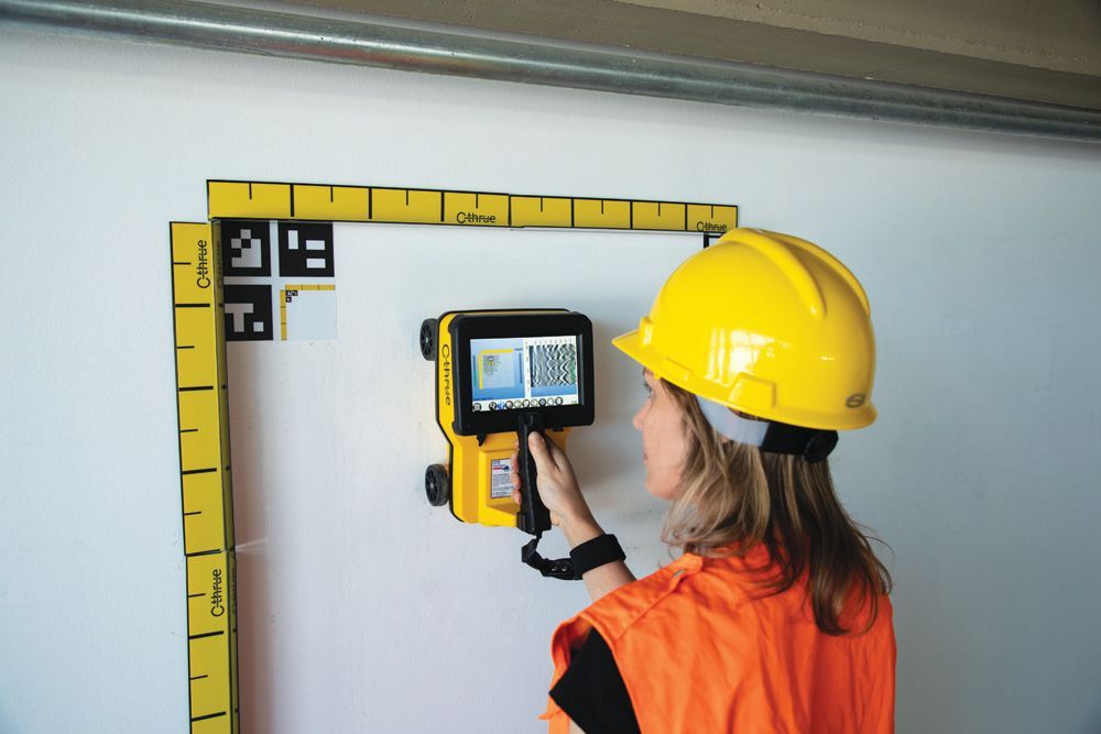 Ein Arbeiter mit gelbem Schutzhelm inspiziert mit einer Wärmebildkamera eine Wandecke.