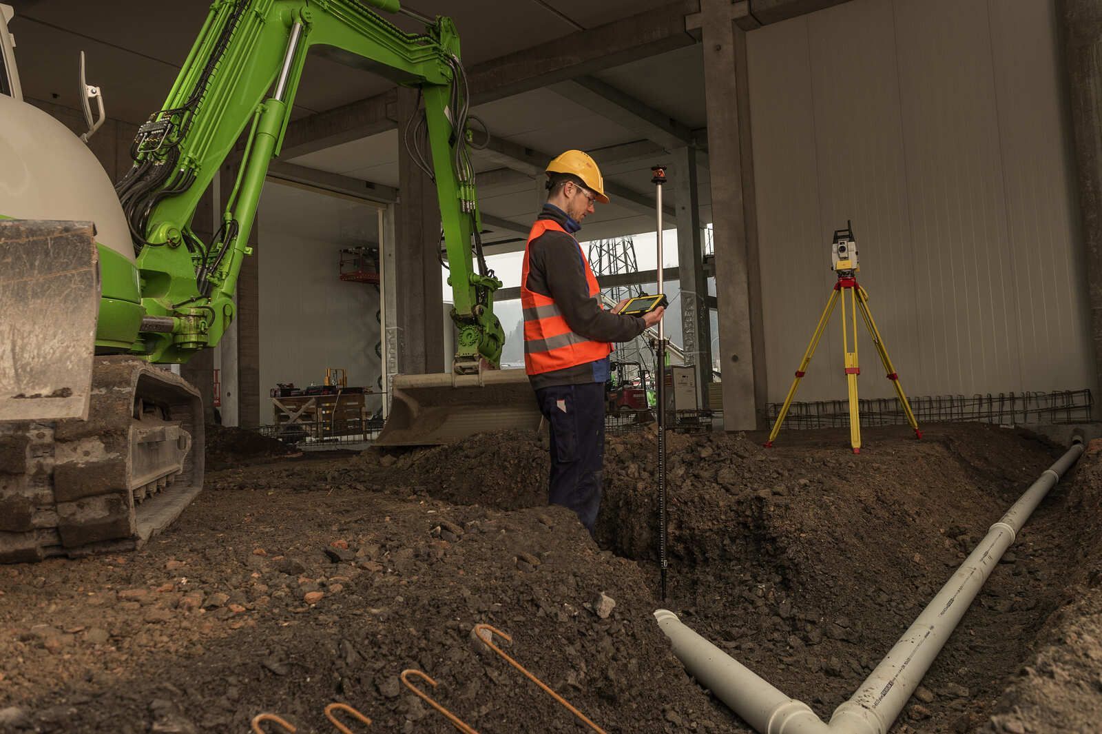 Ein Bauarbeiter steht im Dreck neben einem grünen Bagger.