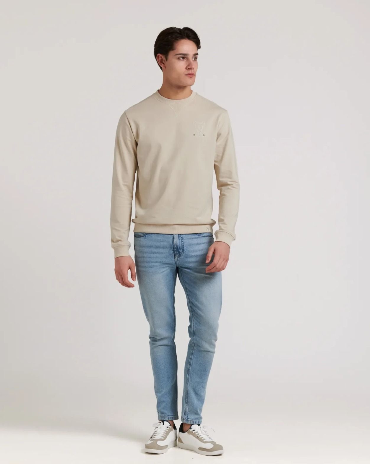 Una persona con una sudadera beige de cuello redondo, vaqueros azul claro y zapatillas deportivas sobre un fondo blanco liso.