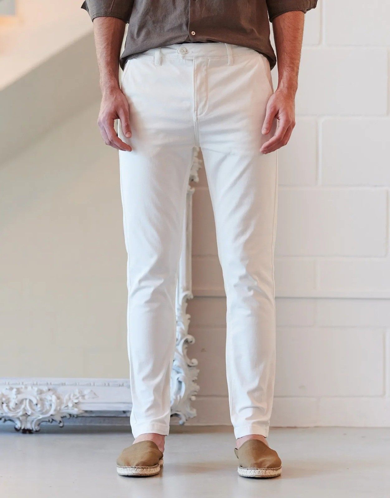 Una persona vestida con una camisa marrón, pantalones blancos y zapatos sin cordones de color canela, de pie en un estudio.