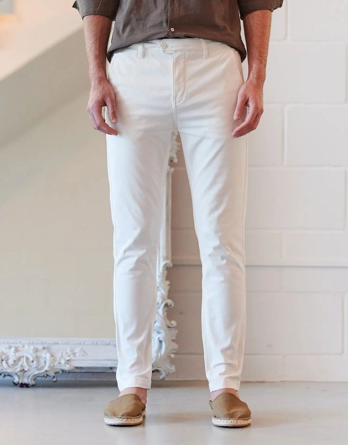 Una persona vestida con una camisa marrón, pantalones blancos y zapatos sin cordones de color canela, de pie en un estudio.