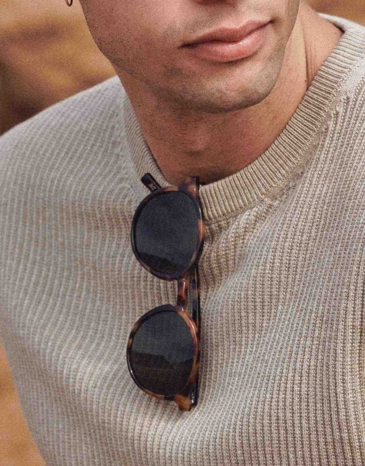 Primer plano de unas gafas de sol de carey que cuelgan del escote de un jersey de punto acanalado de color beige.