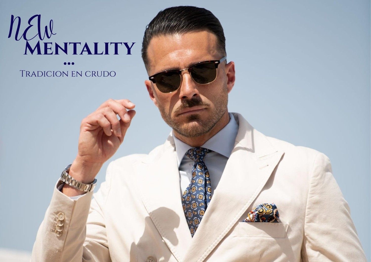 Un hombre con una chaqueta color crema, camisa azul claro y corbata estampada lleva gafas de sol sobre un fondo azul claro.