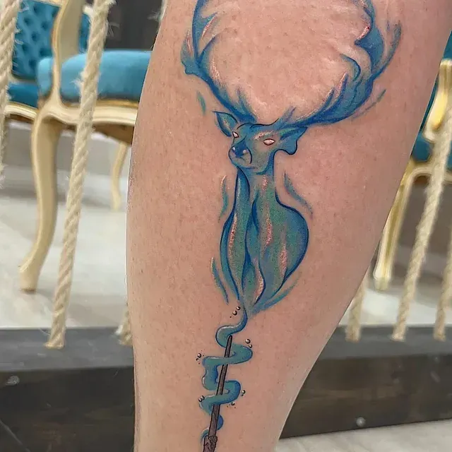 Tatuaje de un ciervo azul con varita en la pierna. El ciervo emana luz, una referencia a Harry Potter.
