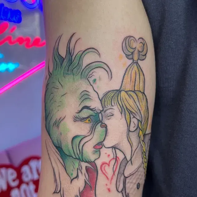 Tatuaje de Grinch besando a Cindy Lou Who; corazón verde, rubio y rojo; en el brazo.
