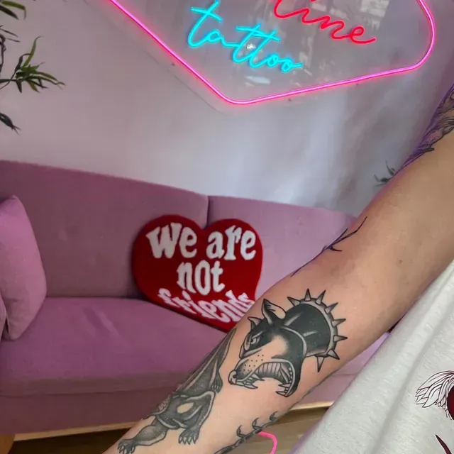 Brazo de una persona con tatuajes, incluyendo un lobo. Sofá rosa, almohada con forma de corazón que dice
