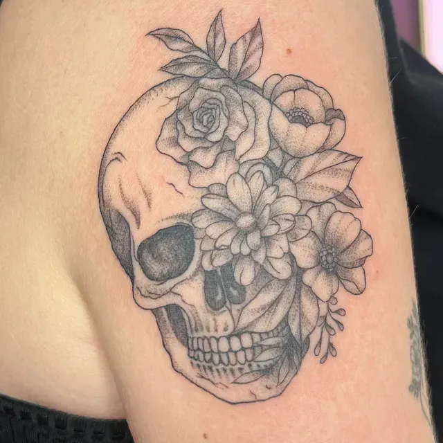 Tatuaje de una calavera con flores floreciendo, entintado en negro en el brazo de una persona.