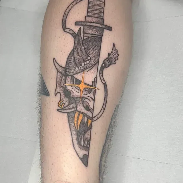Tatuaje de una máscara de demonio bisecada por una daga; dientes naranjas y estrella en el ojo de la máscara.