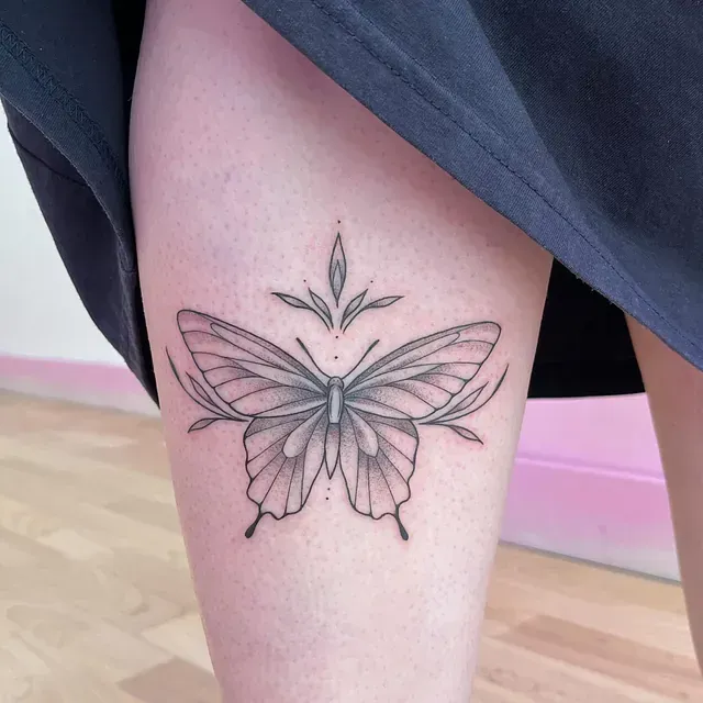 Tatuaje de mariposa en el muslo, con alas detalladas con sombreado, hojas y un diseño central.