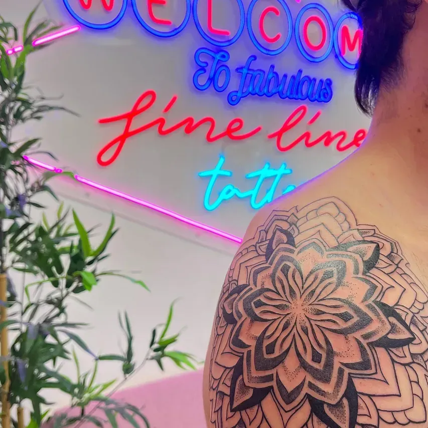 Persona con tatuaje en el hombro posando frente a un letrero de neón en un salón de tatuajes.