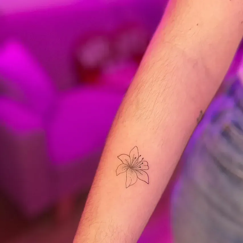 Tatuaje de lirio de líneas finas en el antebrazo de una persona, sobre un fondo morado borroso.