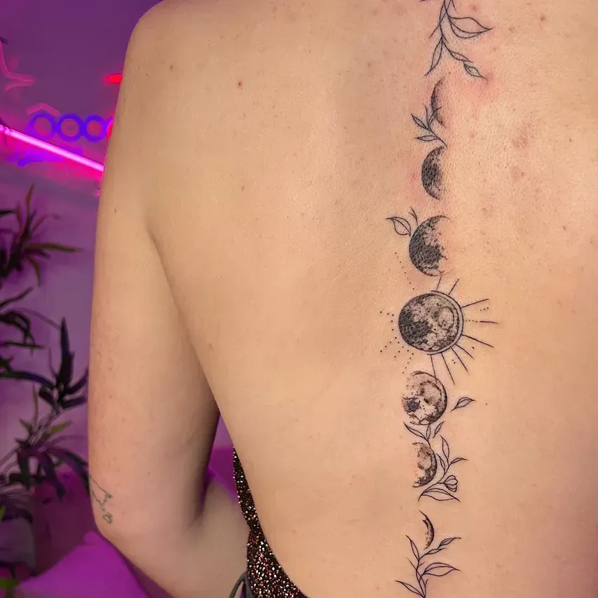 Tatuaje en la espalda: fases de la luna a lo largo de la columna con elementos botánicos en tinta negra.