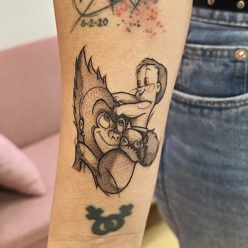 Tatuaje de un gorila de dibujos animados sosteniendo a un niño, con un monito. Hecho en tinta negra en el brazo.