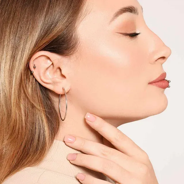 Mujer con pendientes de aro, un piercing en el labio y un dedo tocándose la cara, vista de perfil.