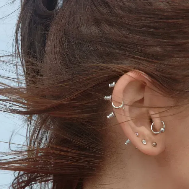 Primer plano de la oreja de una persona con múltiples perforaciones plateadas; cabello castaño enmarcando la oreja.