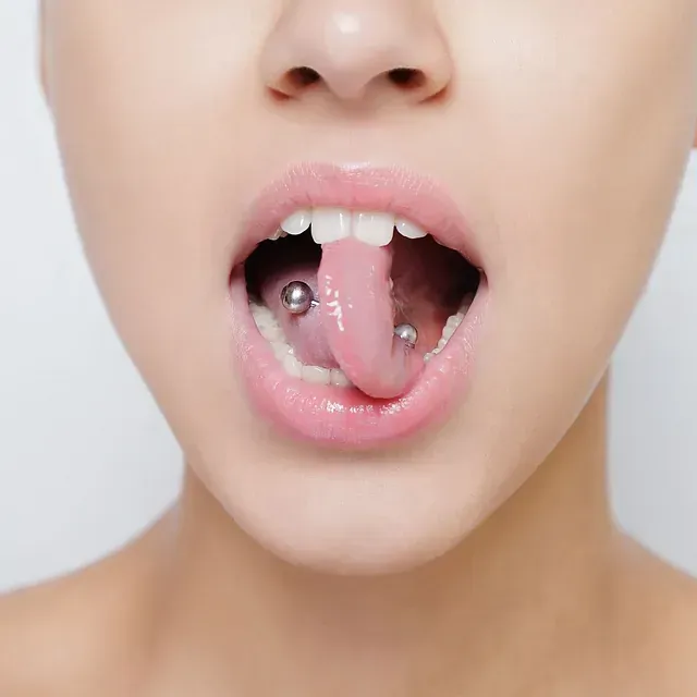 Primer plano de la boca abierta de una persona con piercings en la lengua. La lengua es rosada y la boca está abierta.