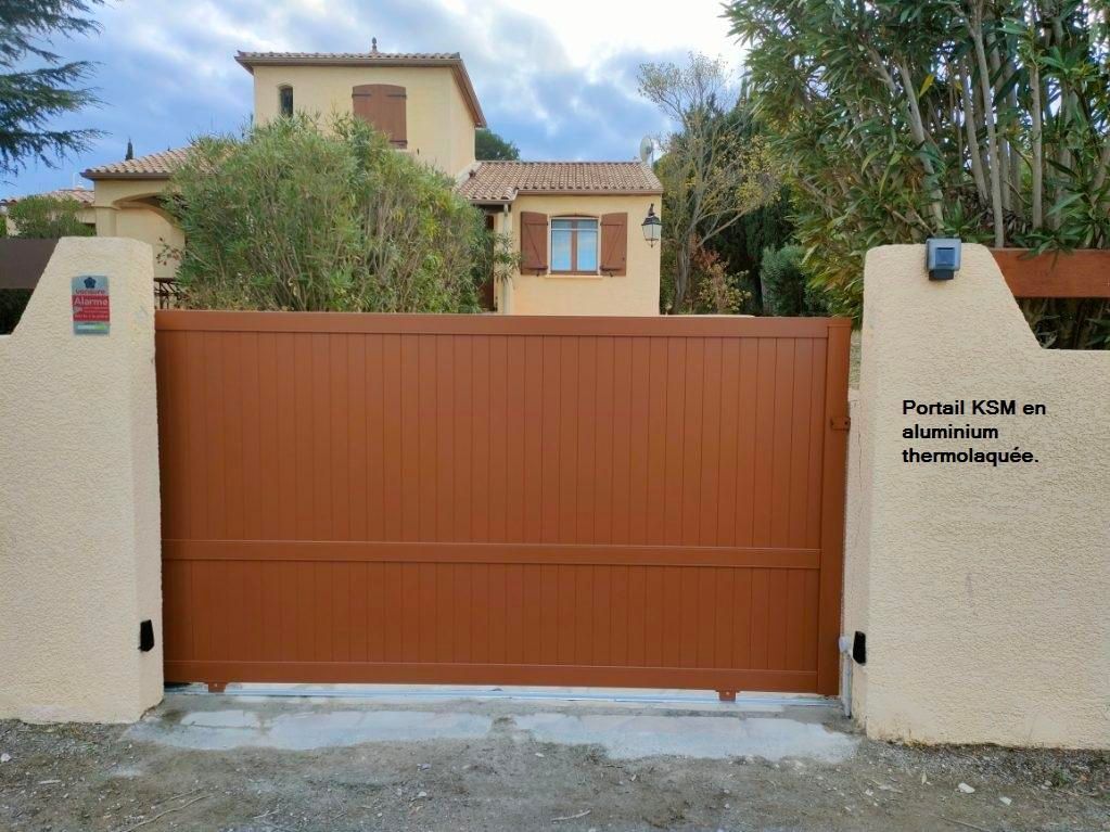 Un portail en métal marron se dresse devant une maison beige, flanqué de piliers beiges.