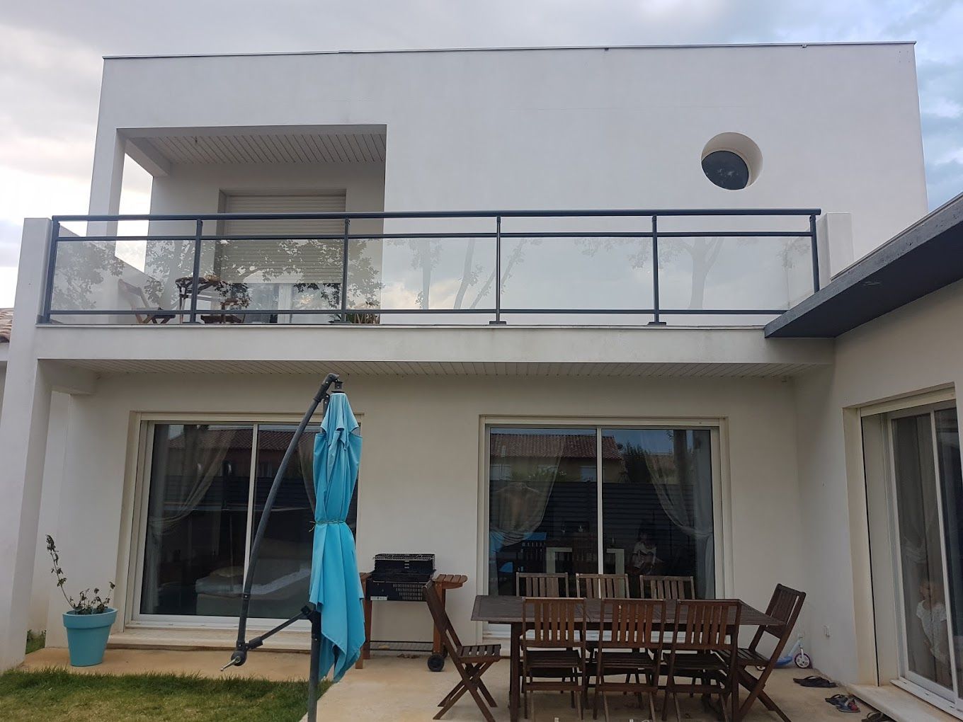 Maison blanche de deux étages avec un balcon à balustrade en verre et de grandes portes coulissantes en verre.