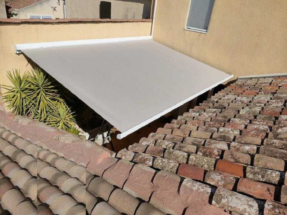 Un auvent blanc ombrage un patio jouxtant un toit de tuiles en terre cuite et un bâtiment beige.
