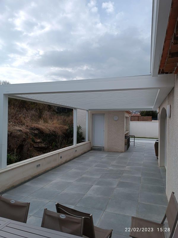 Pergola blanche, sol carrelé gris, murs aux tons neutres et vue sur une colline.