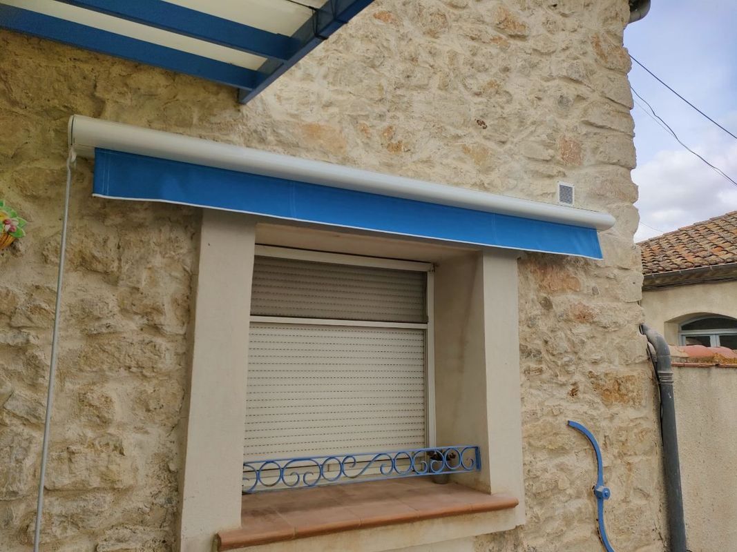 Un auvent bleu surplombe une fenêtre à volet blanc fermé, sur un bâtiment en pierre.