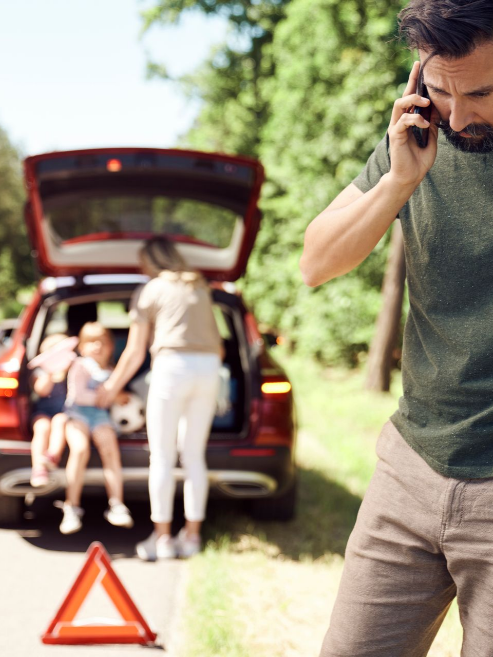 Un homme au téléphone en premier plan avec sa femme et deux enfants assis dans le coffre d'une voiture