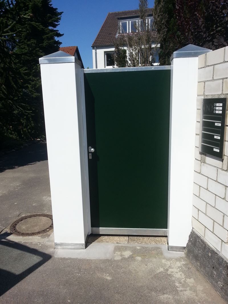 Zwischen zwei weißen Säulen vor einer weißen Backsteinmauer befindet sich ein grünes Tor.